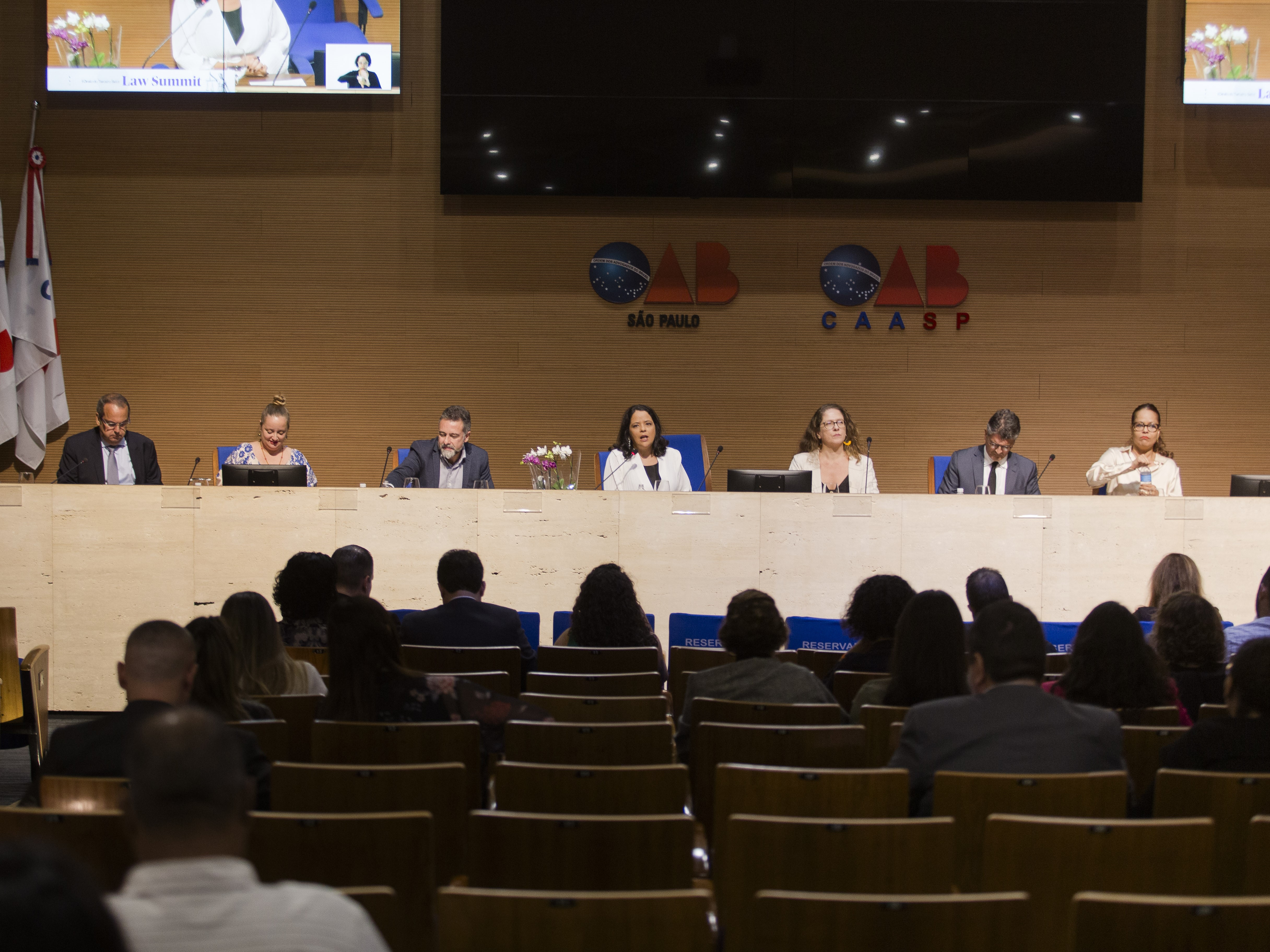 Um grupo de sete pessoas sentadas a frente de uma tela grande, no meio da sala.