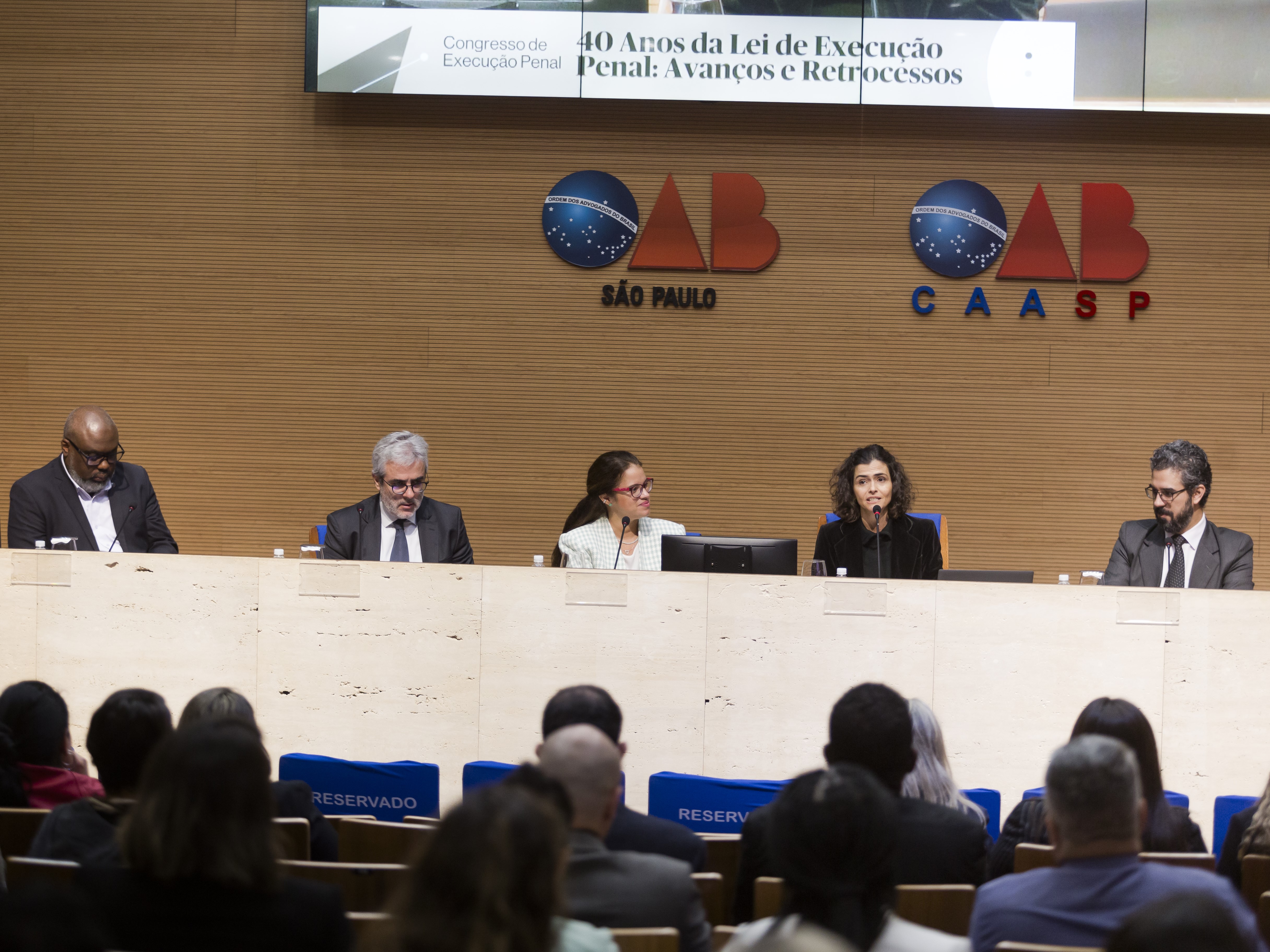 Grupo de cinco pessoas sentadas atrás de um balcão, a frente de uma parede com os símbolos "OAB São Paulo" e "OAB CAASP". Uma plateia assiste a fala das cinco pessoas.