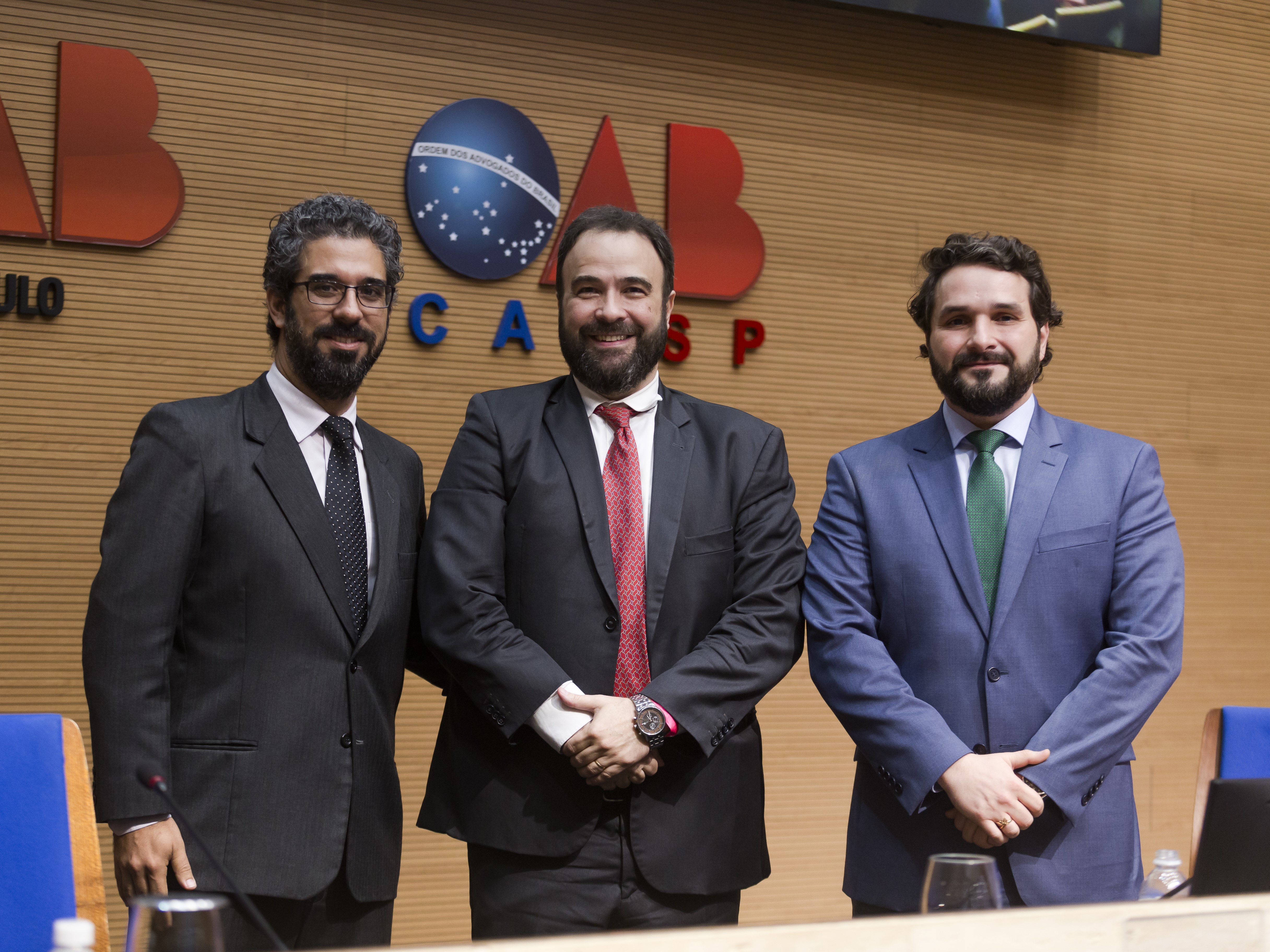 Três homens em pé, vestidos em trajes formais, a frente de uma parede com os símbolos "OAB São Paulo" e "OAB CAASP".