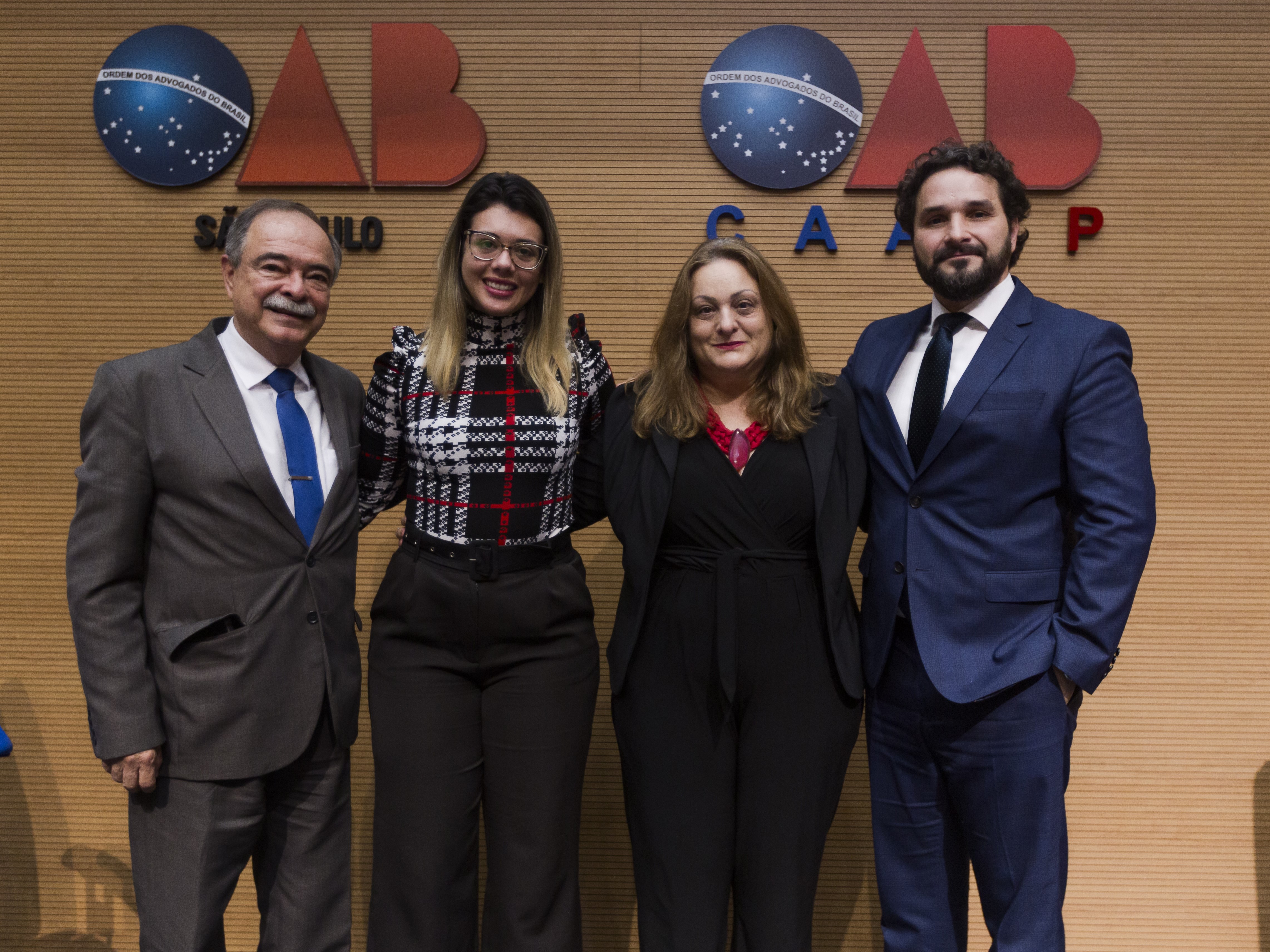 Quatro pessoas posicionadas em pé, vestidas em trajes formais, a frente de uma parede de madeira com os símbolos "OAB São Paulo" e "OAB CAASP"