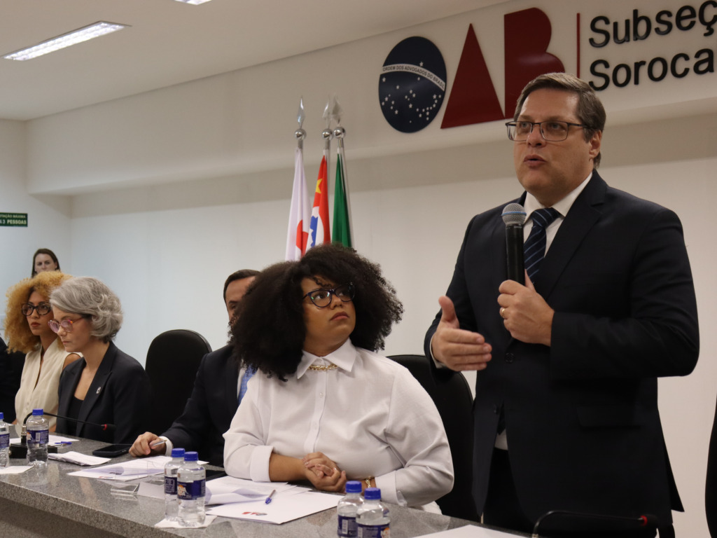 homem branco e óculos discursa em pé com microfone em meio a plateia
